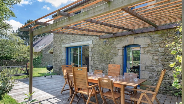 Een houten terras met een pergola, stenen muren en een eettafel.