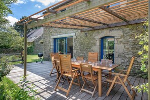 Une terrasse en bois agrémentée d’une pergola, de murs en pierre et d’une table dressée pour le repas.