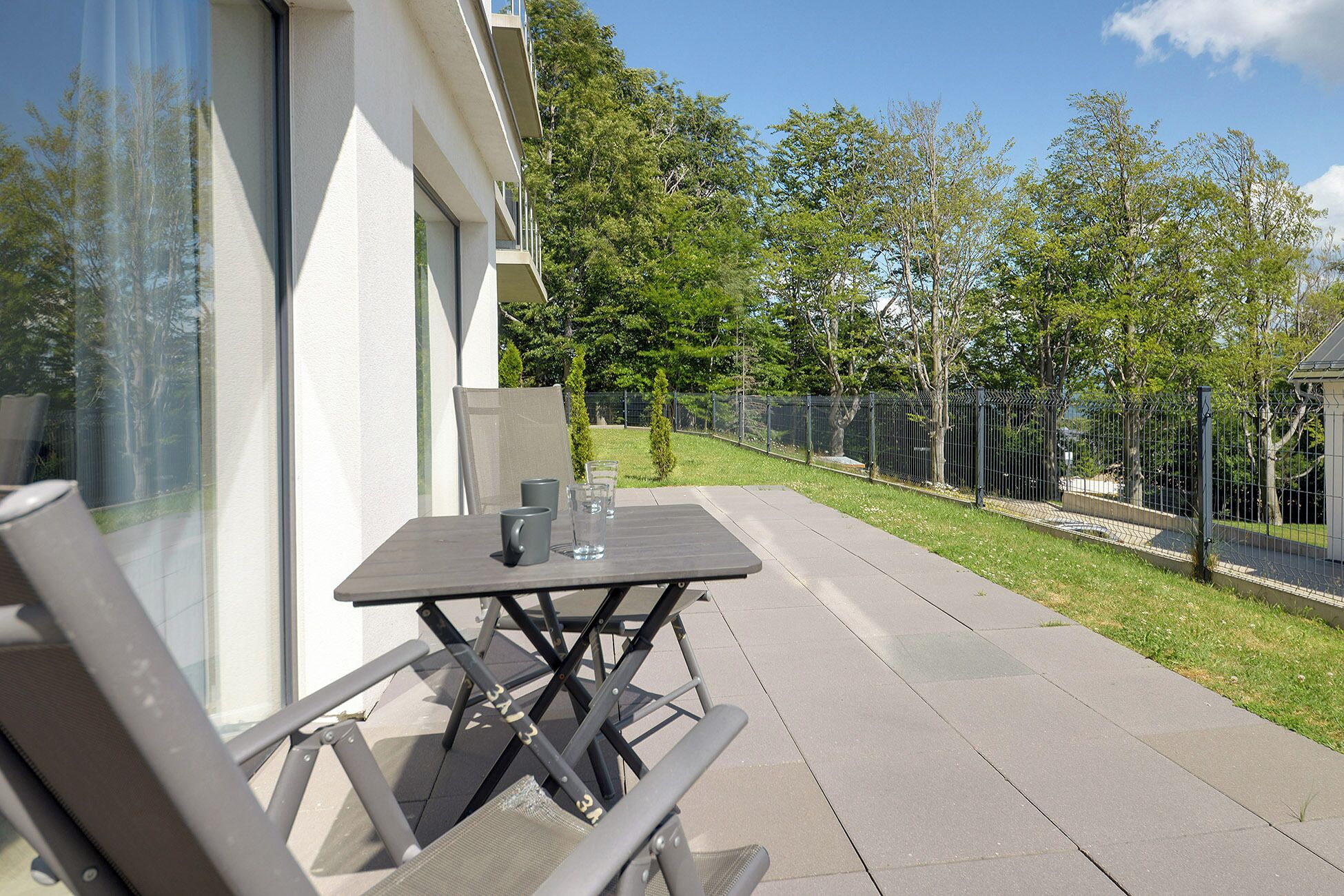 Een terras buiten met een tafel, stoelen, een glas water en uitzicht op een omheind tuintje.