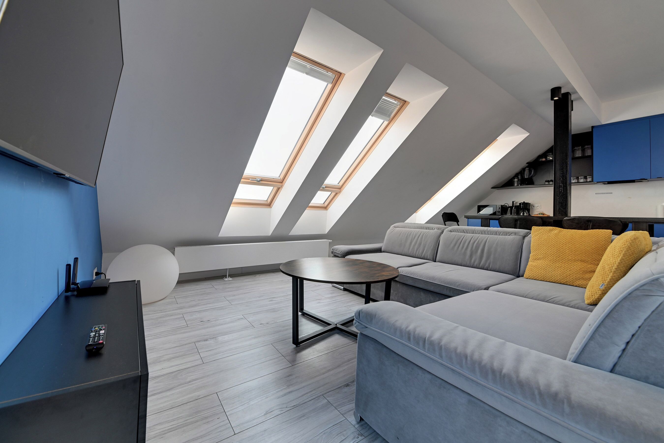En modern lägenhet i loftstil med en stor grå hörnsoffa, ett litet runt bord och ett köksområde med blå skåp.