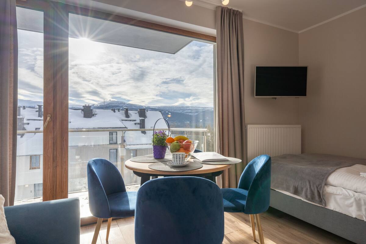 A modern hotel room with a large window, a bed, a TV, and a dining area with chairs and a table.