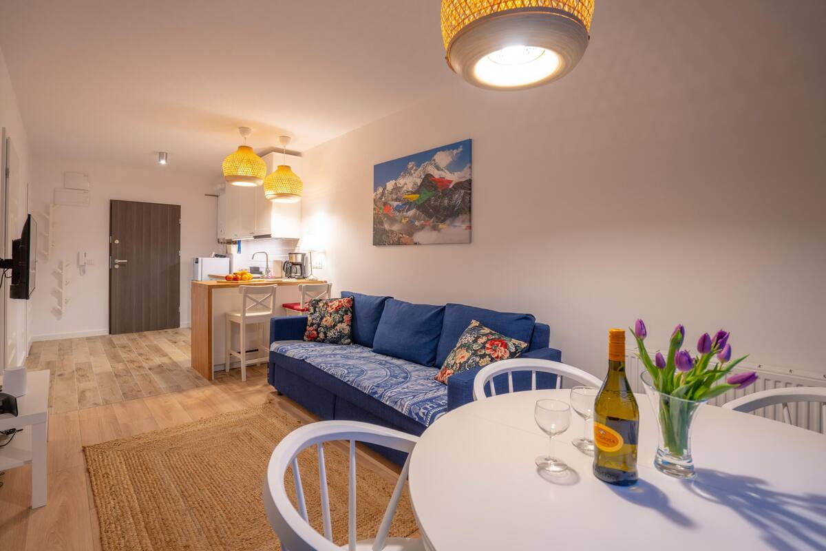 A modern living room with a blue sofa, white chairs, a dining table with wine glasses, and a kitchen in the background.