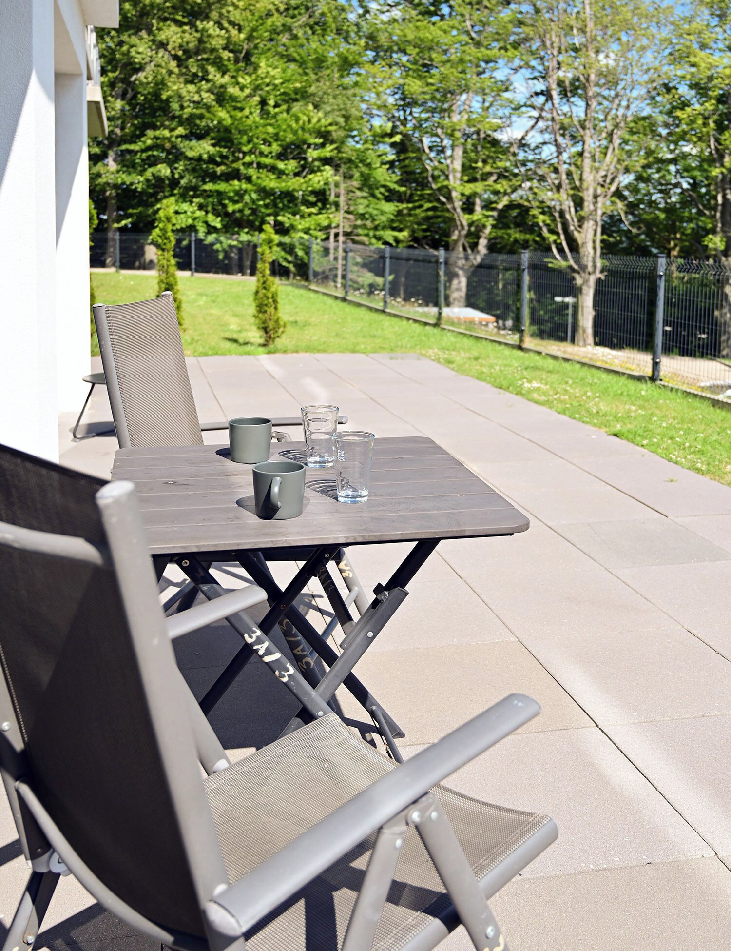 Un patio al aire libre con una mesa y sillas, una mesita con una taza y un vaso, y vista a un patio cercado con árboles.