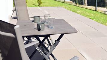 Un patio al aire libre con una mesa y sillas, una mesita con una taza y un vaso, y vista a un patio cercado con árboles.