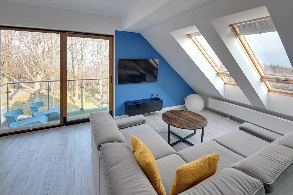 A modern loft bedroom with a large window, a flat-screen TV, and a sofa with cushions.