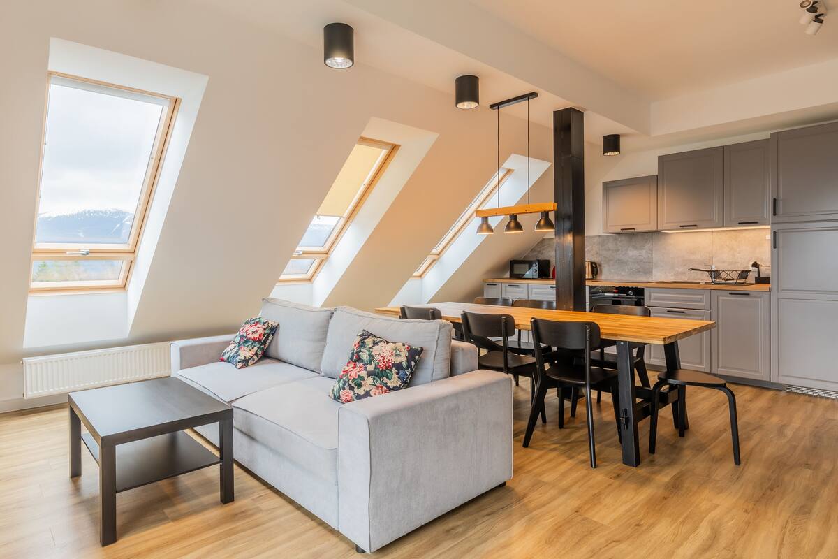 A modern loft apartment with a dining area, kitchen, and a gray sofa with floral cushions.