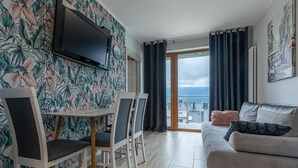 A living room with a sofa, a TV mounted on the wall, a dining table with chairs, and a view of the sea through large windows. - RentPlanet - Apartament Schroniskowa (Szklarska Poreba)