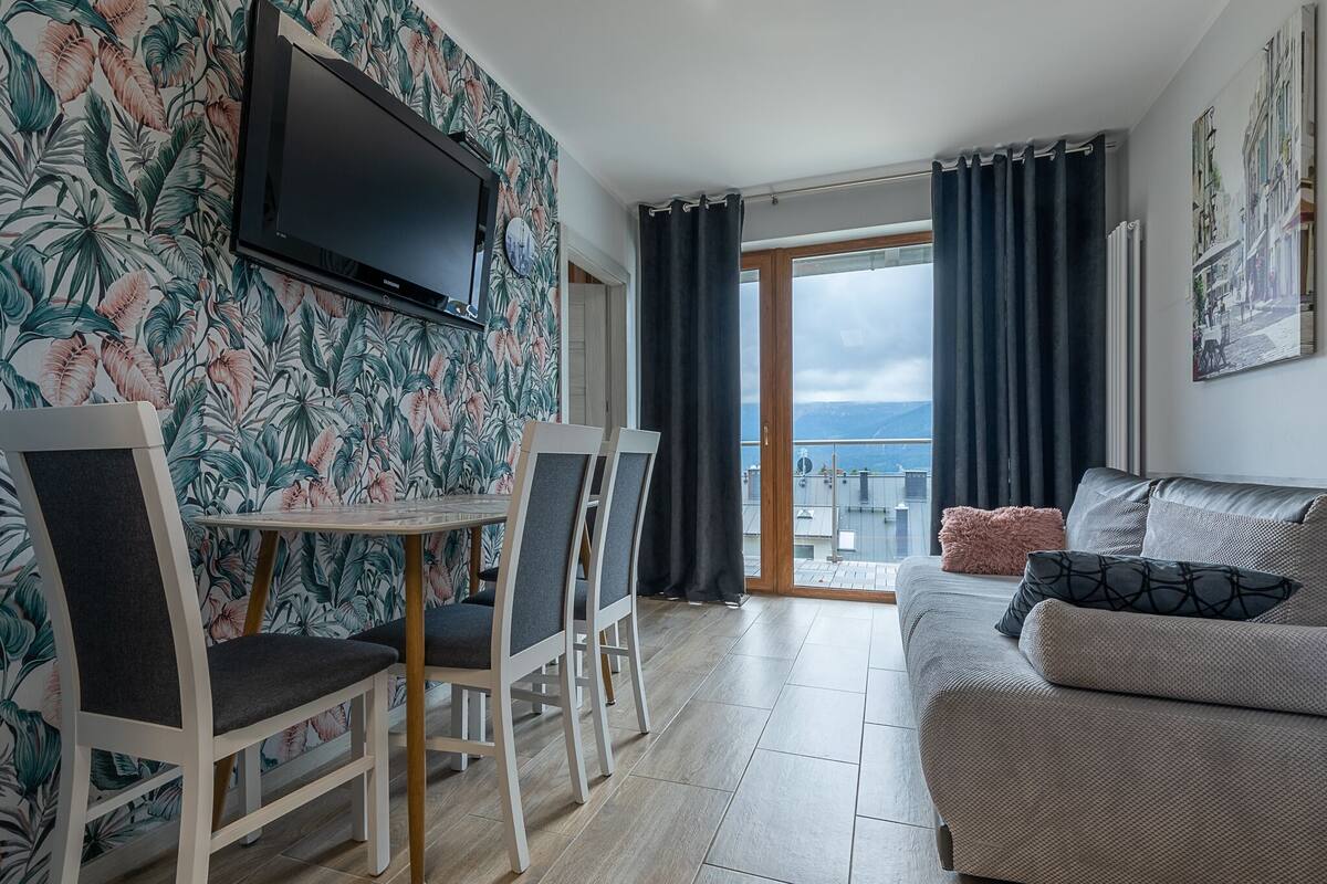 A living room with a sofa, a TV mounted on the wall, a dining table with chairs, and a view of the sea through large windows.