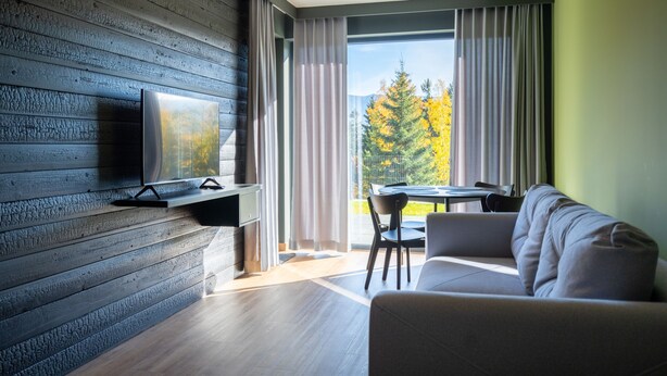 A modern living room with a flat-screen TV mounted on a wooden wall, a sofa, a dining table with chairs, and a large window with curtains. - RentPlanet - Apartament Schroniskowa (Szklarska Poreba)