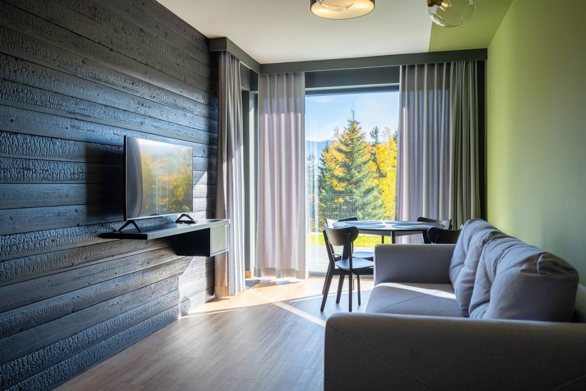 A modern living room with a flat-screen TV mounted on a wooden wall, a sofa, a dining table with chairs, and a large window with curtains.