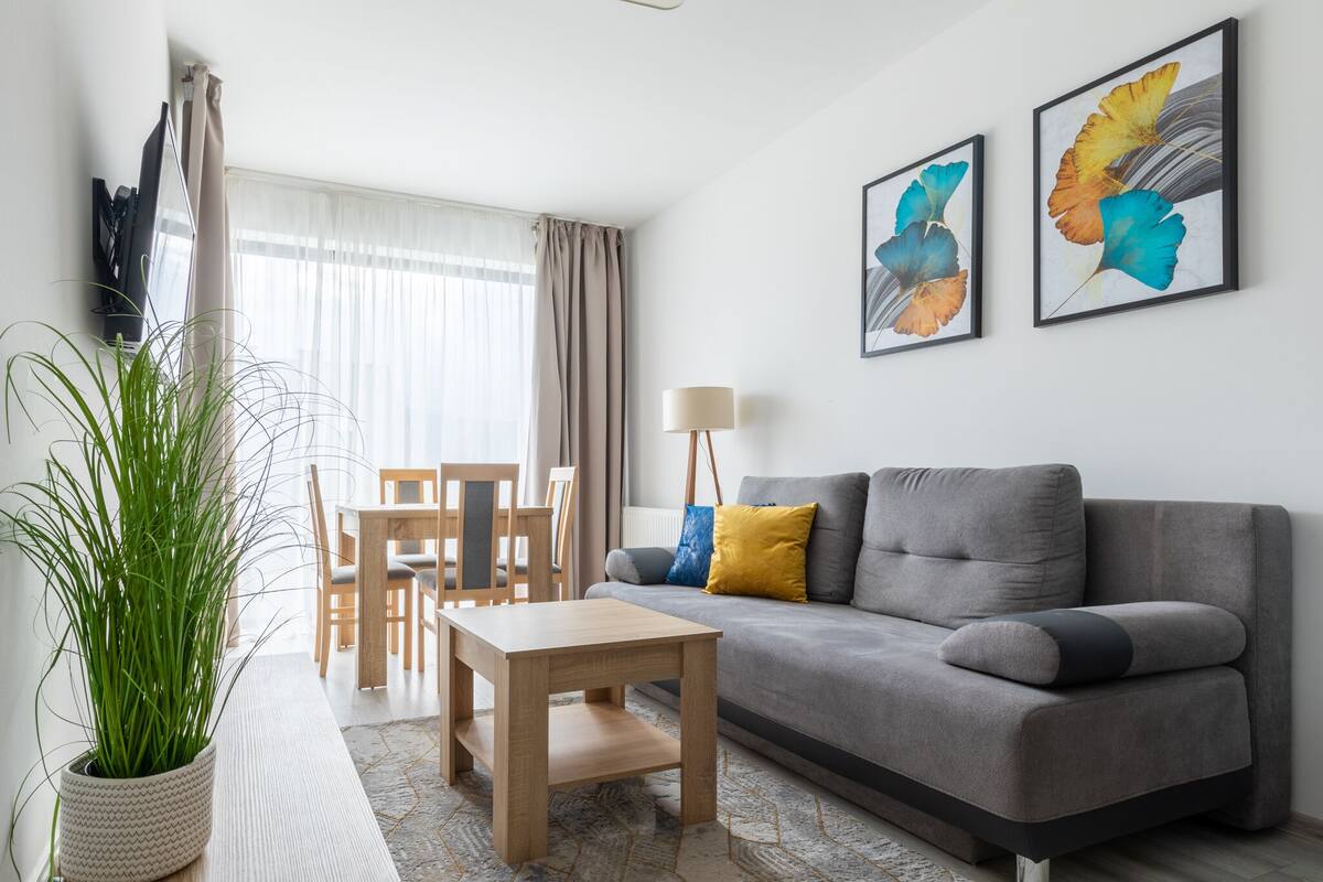 A modern living room with a grey sofa, a wooden coffee table, and a dining area with a table and chairs. There is a large potted plant and two framed artworks on the wall.