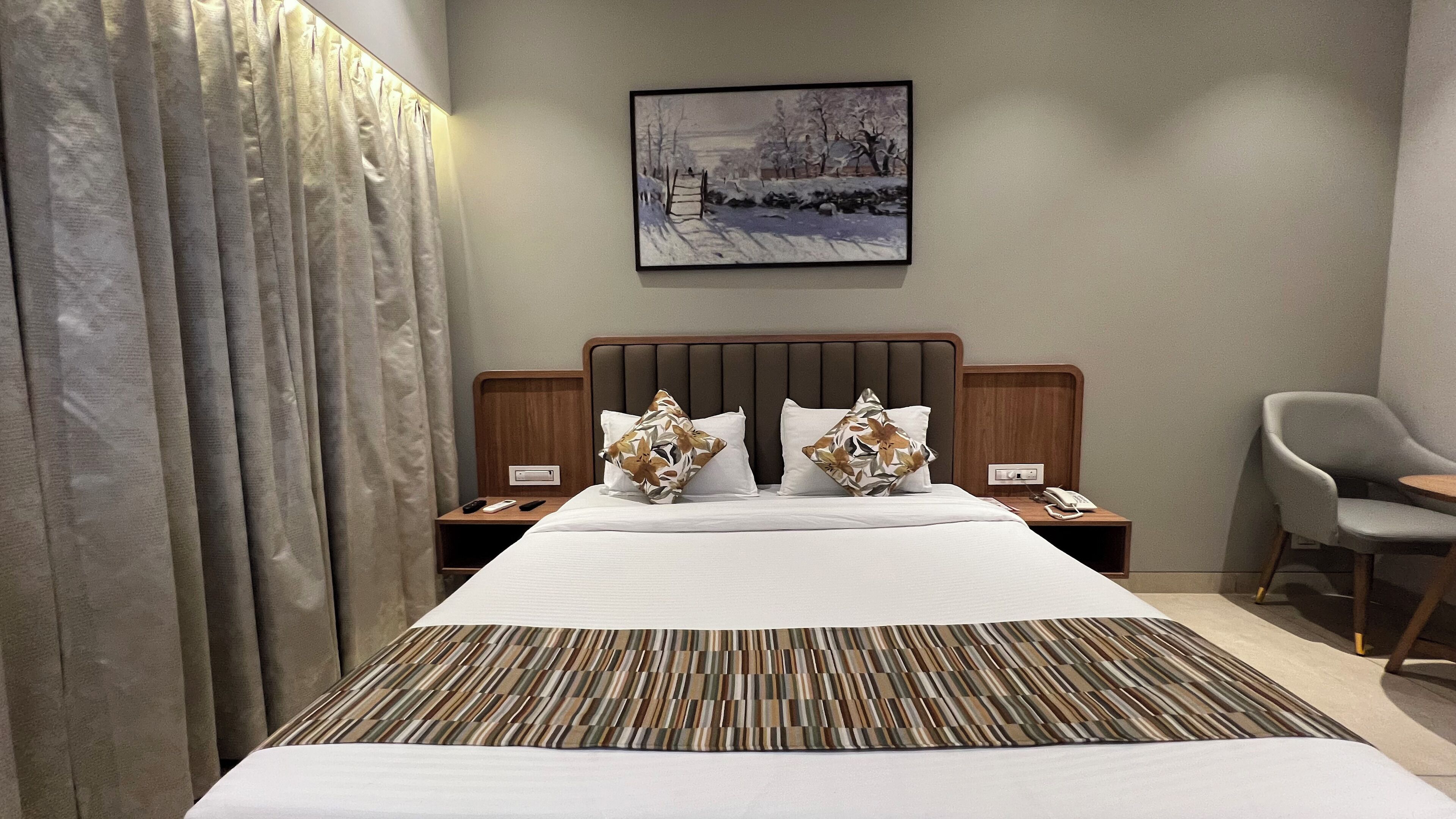 Ein Hotelzimmer mit einem großen Bett, einem Kopfteil, zwei Nachttischen mit Telefonen, einem Sessel und einem Gemälde an der Wand.