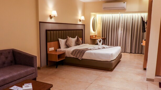 A hotel room with a bed, a sofa, a nightstand, a lamp, and a curtain. - Rudra Shelter Business Hotel (Vasai)