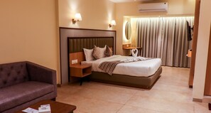 A hotel room with a bed, a sofa, a nightstand, a lamp, and a curtain. - Rudra Shelter Business Hotel (Vasai)