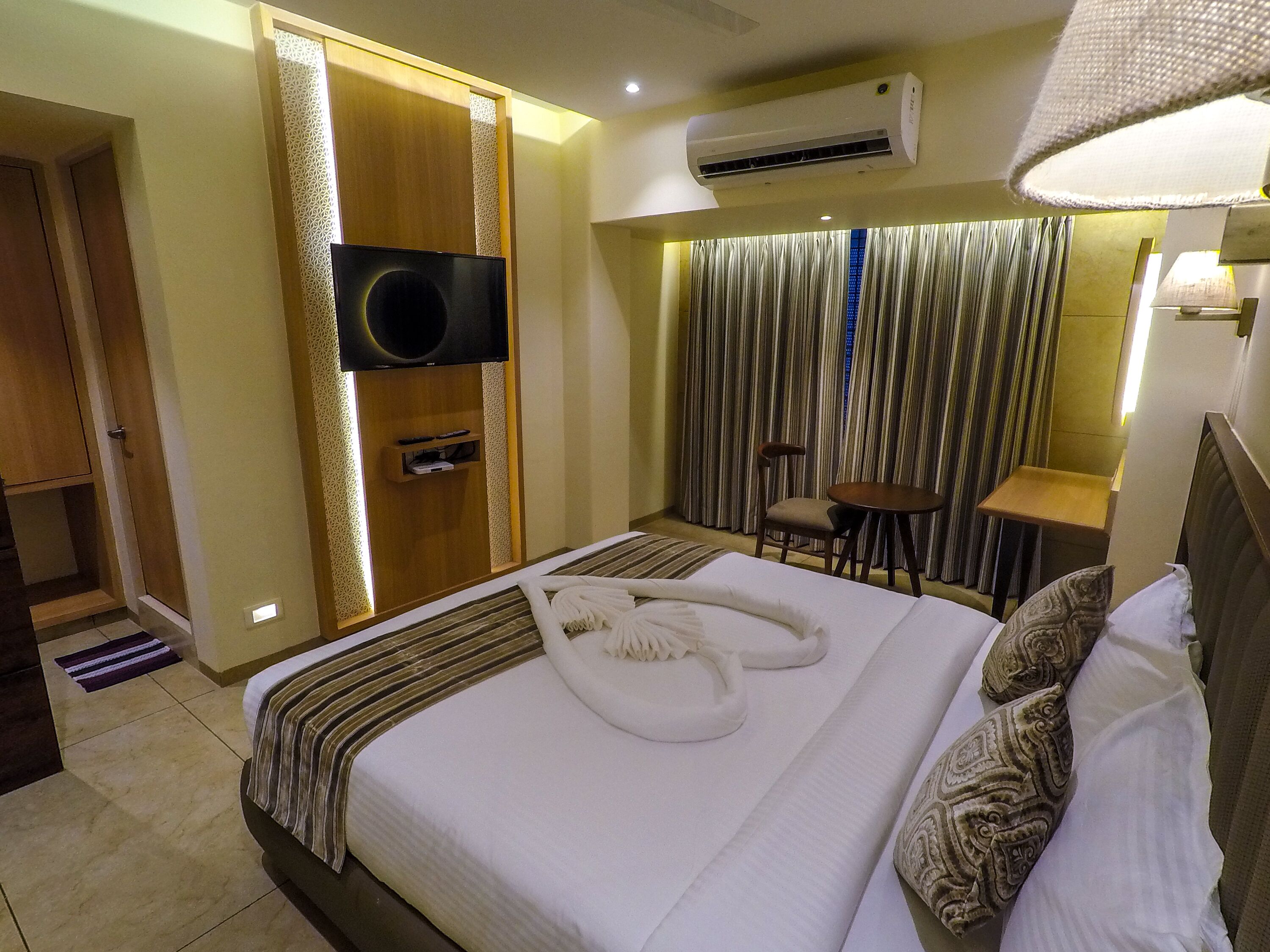 A hotel room with a bed, a TV mounted on a wooden panel, a desk with a chair, and an air conditioning unit.