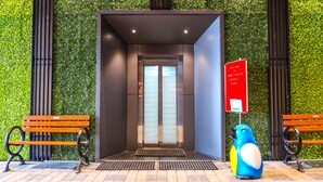 An elevator entrance with a green wall, two wooden benches, and a blue penguin statue. - Rudra Shelter Business Hotel (Vasai)