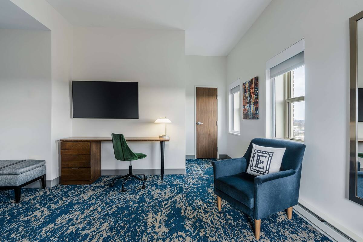A modern hotel room with a large flat-screen TV, a desk with a chair, a blue armchair, a large mirror, and a patterned carpet.