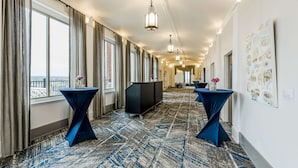 Corredor de hotel com grandes janelas, mesas cobertas com tecido azul e obras de arte nas paredes.