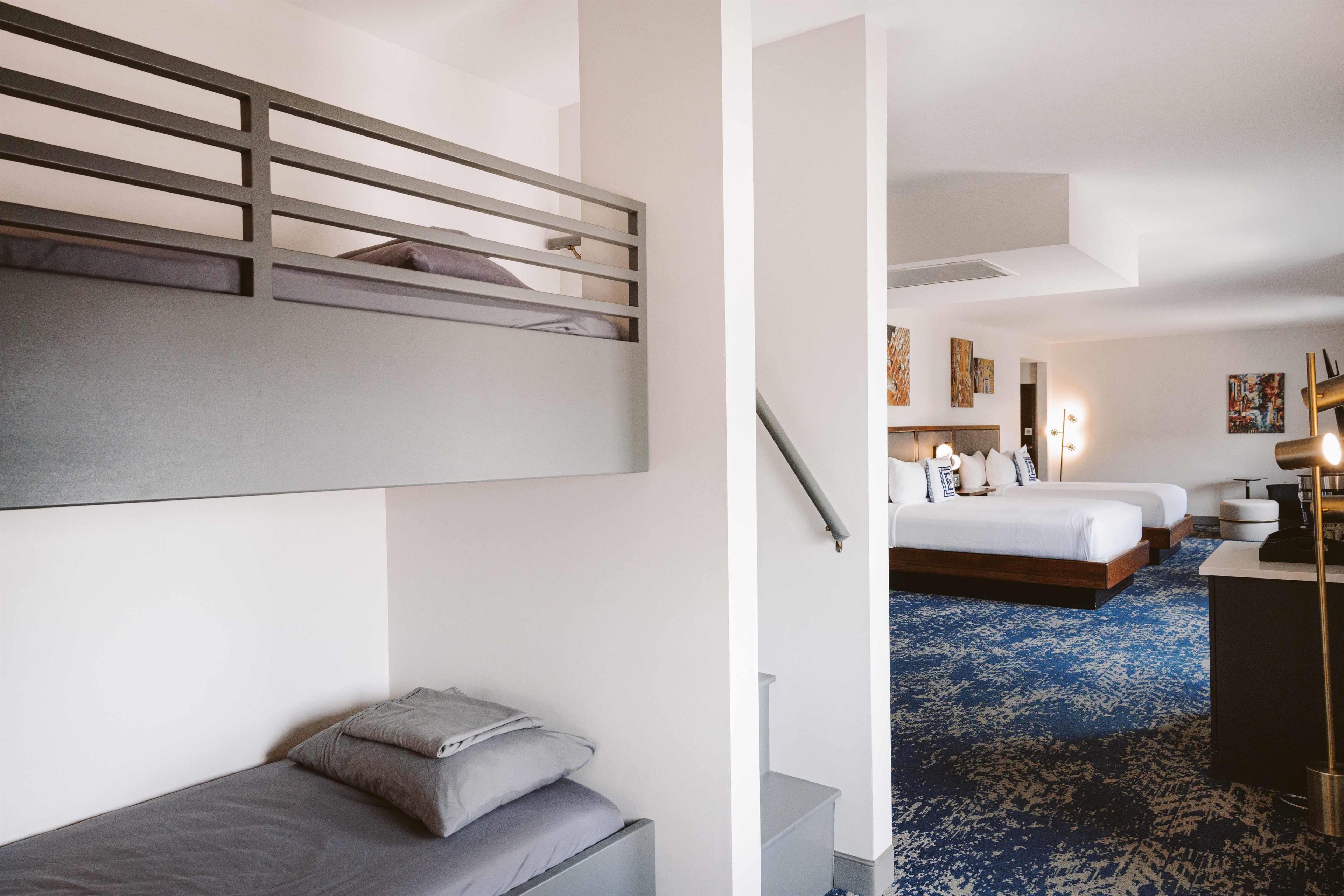 A hotel room with a bunk bed, a single bed, a desk with a lamp, and a blue patterned carpet.