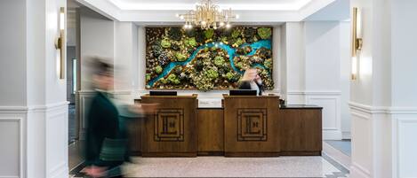 A hotel reception area with a modern chandelier, a large wall tapestry, and a reception desk.