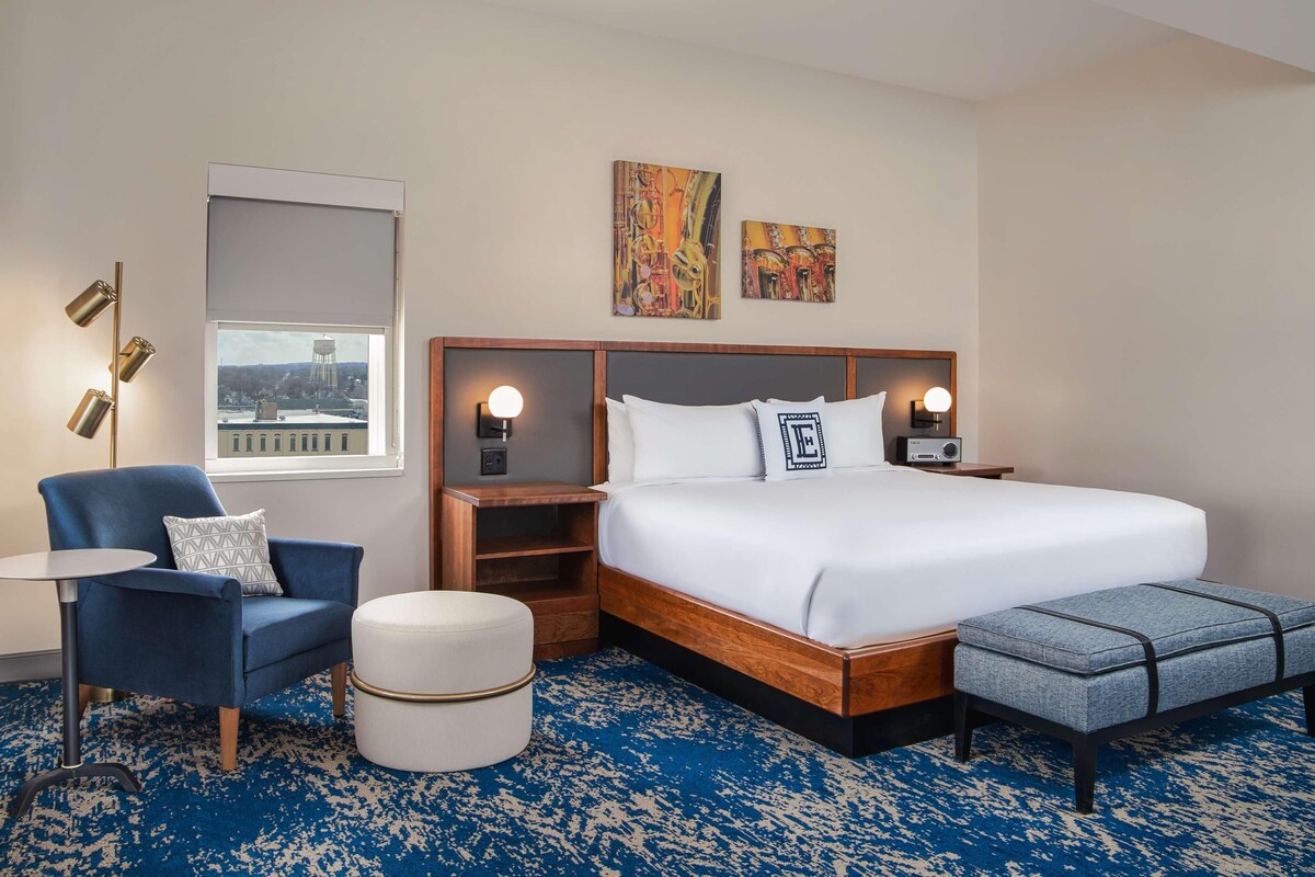 A hotel room with a large bed, a chair, a small table, a round ottoman, and a window with a view of a water tower.