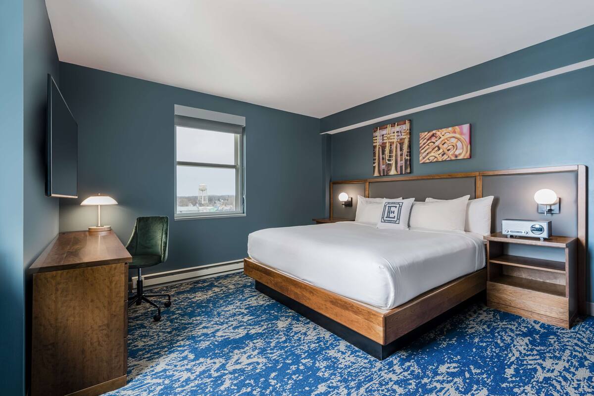 A bedroom with a large bed, a desk, a chair, a TV, and artwork on the wall.