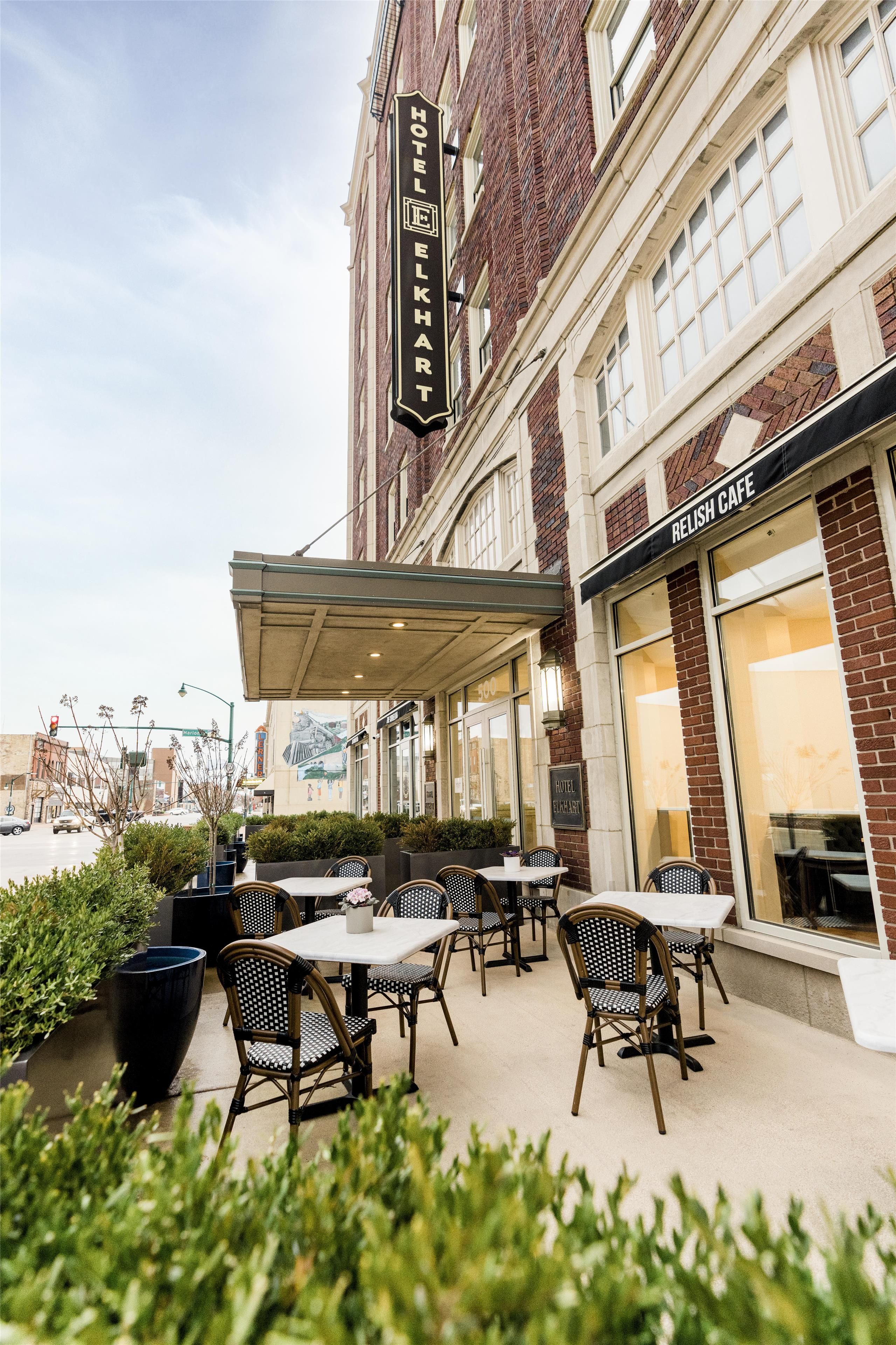 An outdoor seating area with tables and chairs, a building with a 'Hotel Elkhart' sign, and a 'Relish Cafe' across the street.