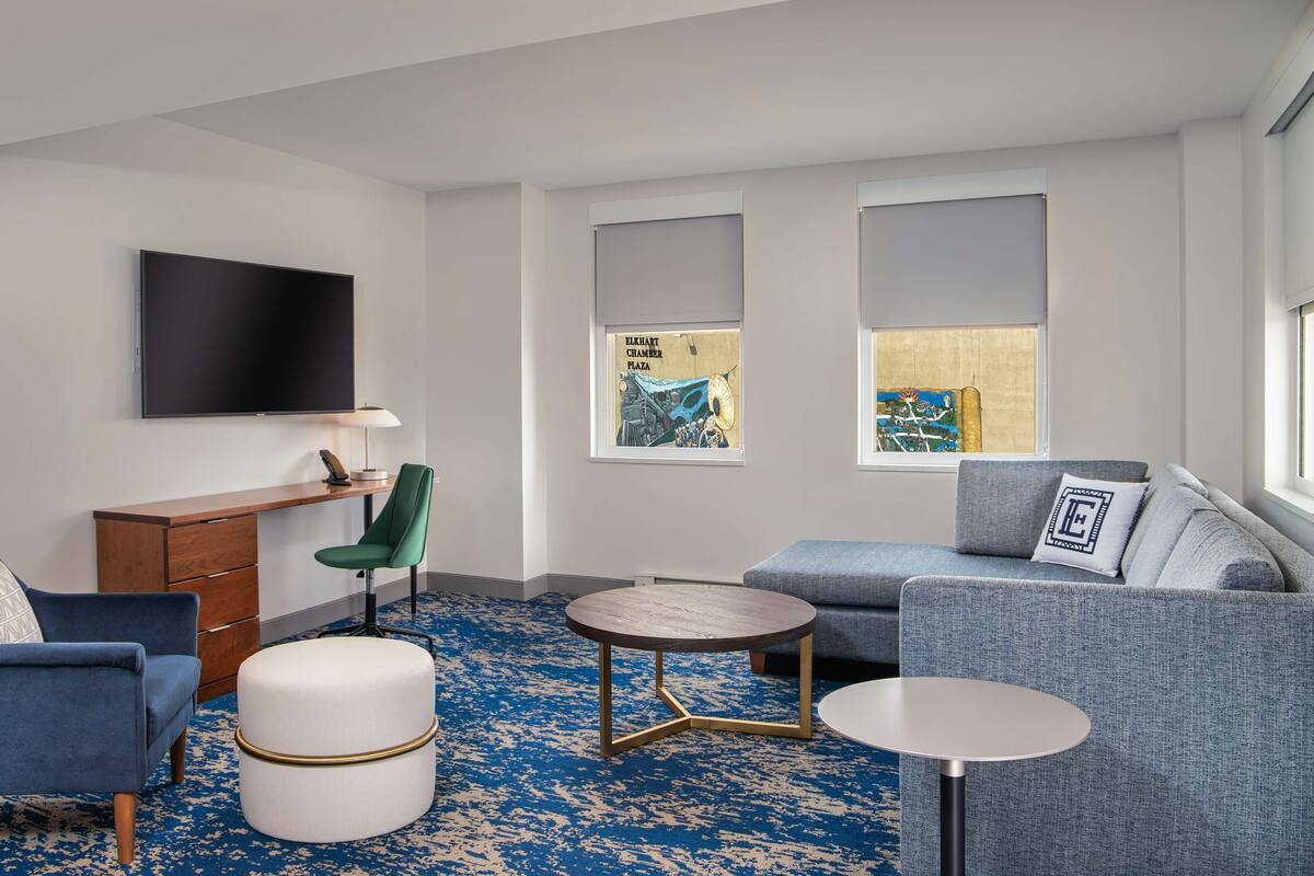 A modern hotel room with a flat-screen TV, a desk with a chair, a sofa, a round coffee table, and a patterned carpet.