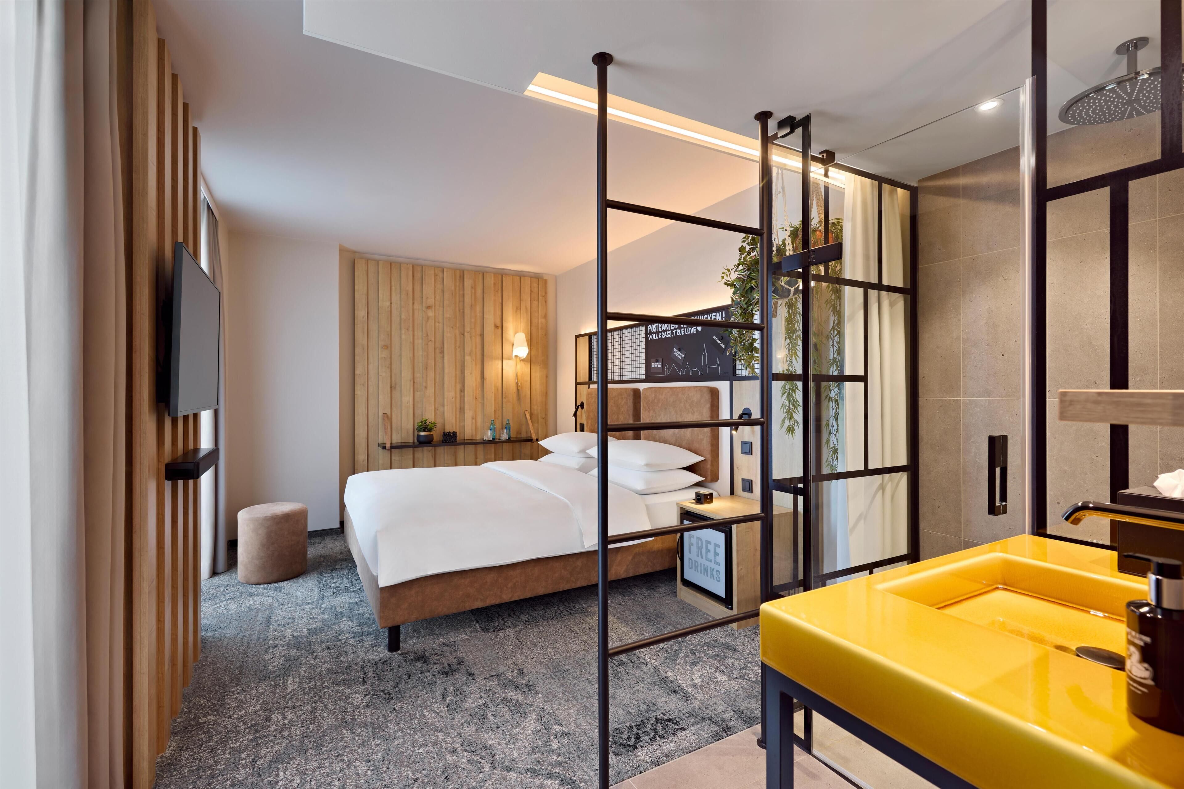 Een moderne hotelkamer met een bed, een geel wastafelmeubel, een douche en een houten scheidingswand.