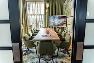 Un salón de conferencias con una mesa larga, sillas verdes, una pantalla de televisión y vistas a la ciudad.