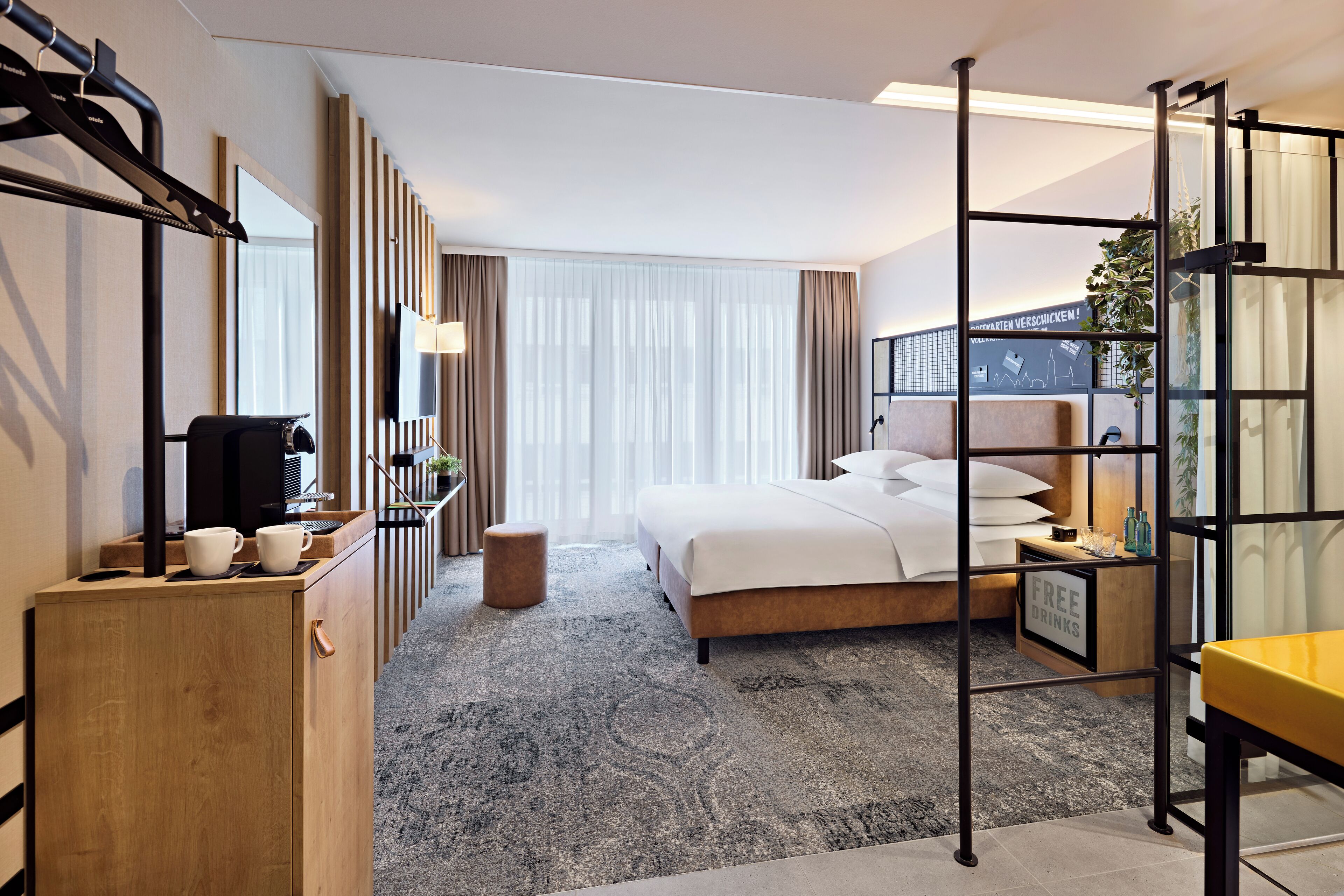 A hotel room with a bed, a desk, a chair, a small table, a mirror, a plant, and a sign offering free drinks.