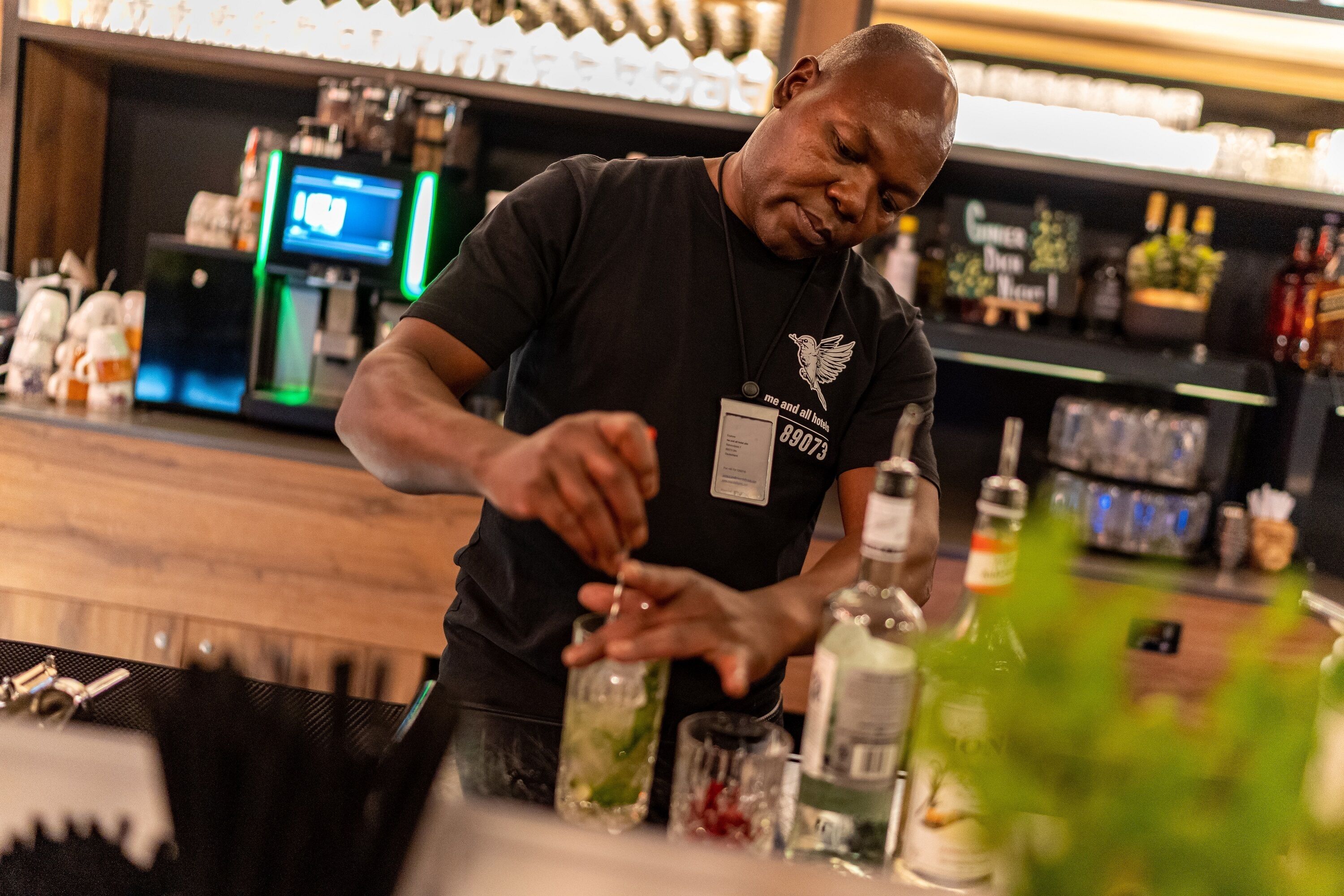 A bartender preparing a drink at a bar with a digital screen and various bottles in the background.
