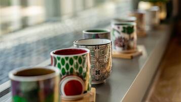 Una hilera de tazas decorativas sobre posavasos de madera junto a una ventana con persianas verticales.