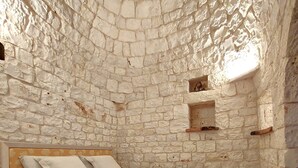 A bedroom with a stone wall, a bed with a wooden headboard, and a small shelf. - Trullo Degli Ulivi (Castellana Grotte)