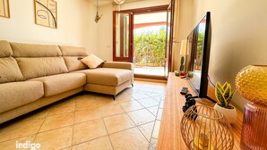 A living room with a beige sectional sofa, a flat-screen TV, a wooden coffee table, and a large window with a view of greenery.