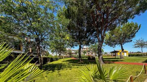 A well-maintained garden with manicured lawns and tropical plants, a paved pathway, and a building with a yellow facade in the background.