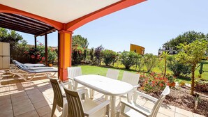 Eine überdachte Terrasse mit Tisch und Stühlen, ein Poolbereich mit Liegestühlen sowie ein Blick auf einen Garten mit verschiedenen Pflanzen und einem Gebäude im Hintergrund.