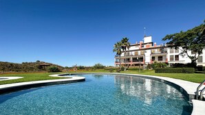 Ein klarer, blauer Pool mit geschwungener Kante, umgeben von gepflegtem Rasen und Palmen. Im Hintergrund ein großes Gebäude mit mehreren Balkonen und einem roten Dach.