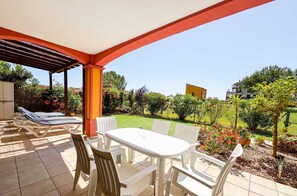 A covered patio with a table and chairs, a pool area with lounge chairs, and a view of a garden with various plants and a building in the background.