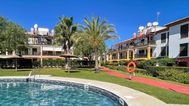A swimming pool with a tiled edge, surrounded by a well-maintained lawn and palm trees. - Apartment with garden & direct access to the pool CAP01 (Ayamonte)