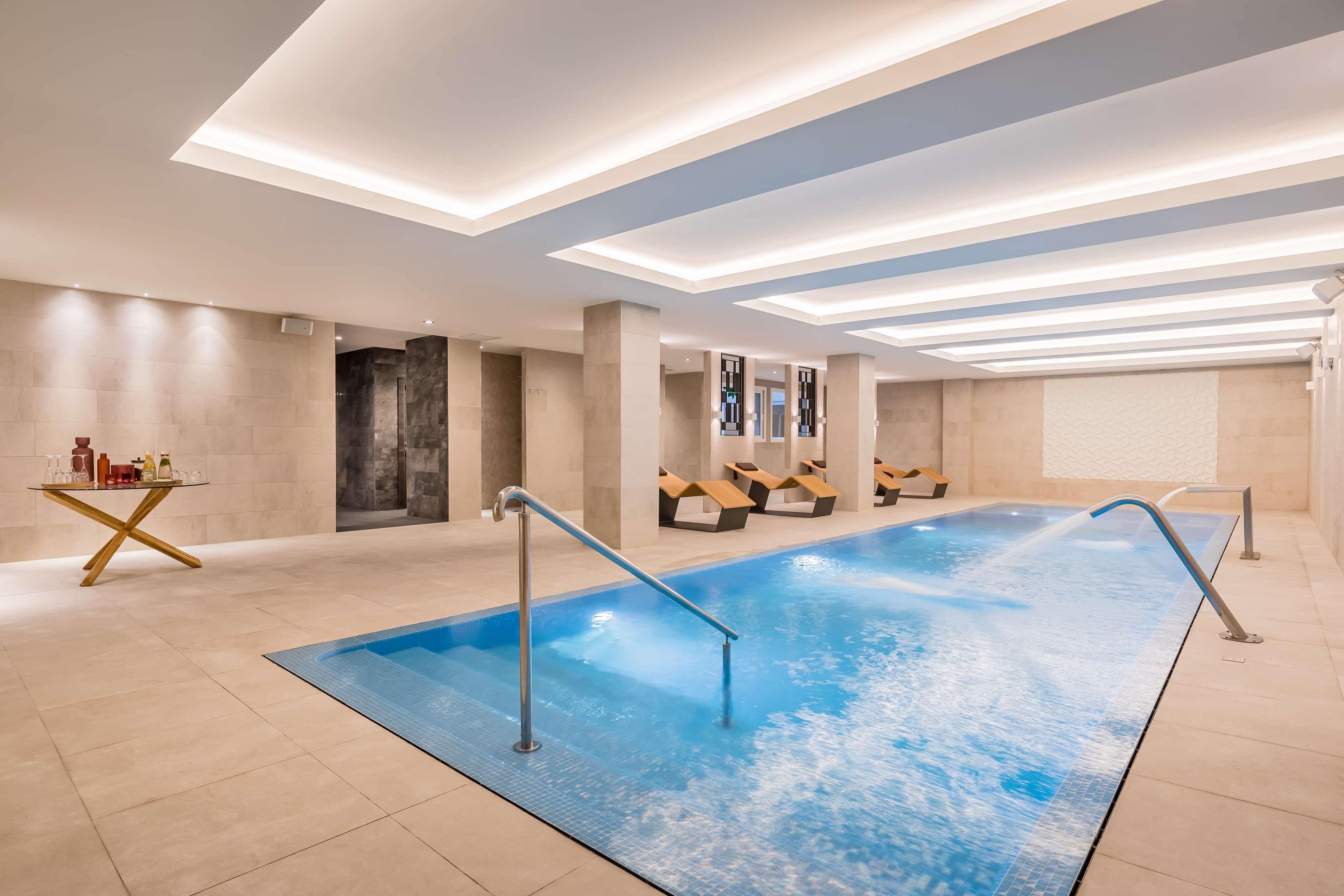 A modern indoor swimming pool with a blue water feature, a glass table with bottles, and a seating area with wooden loungers.