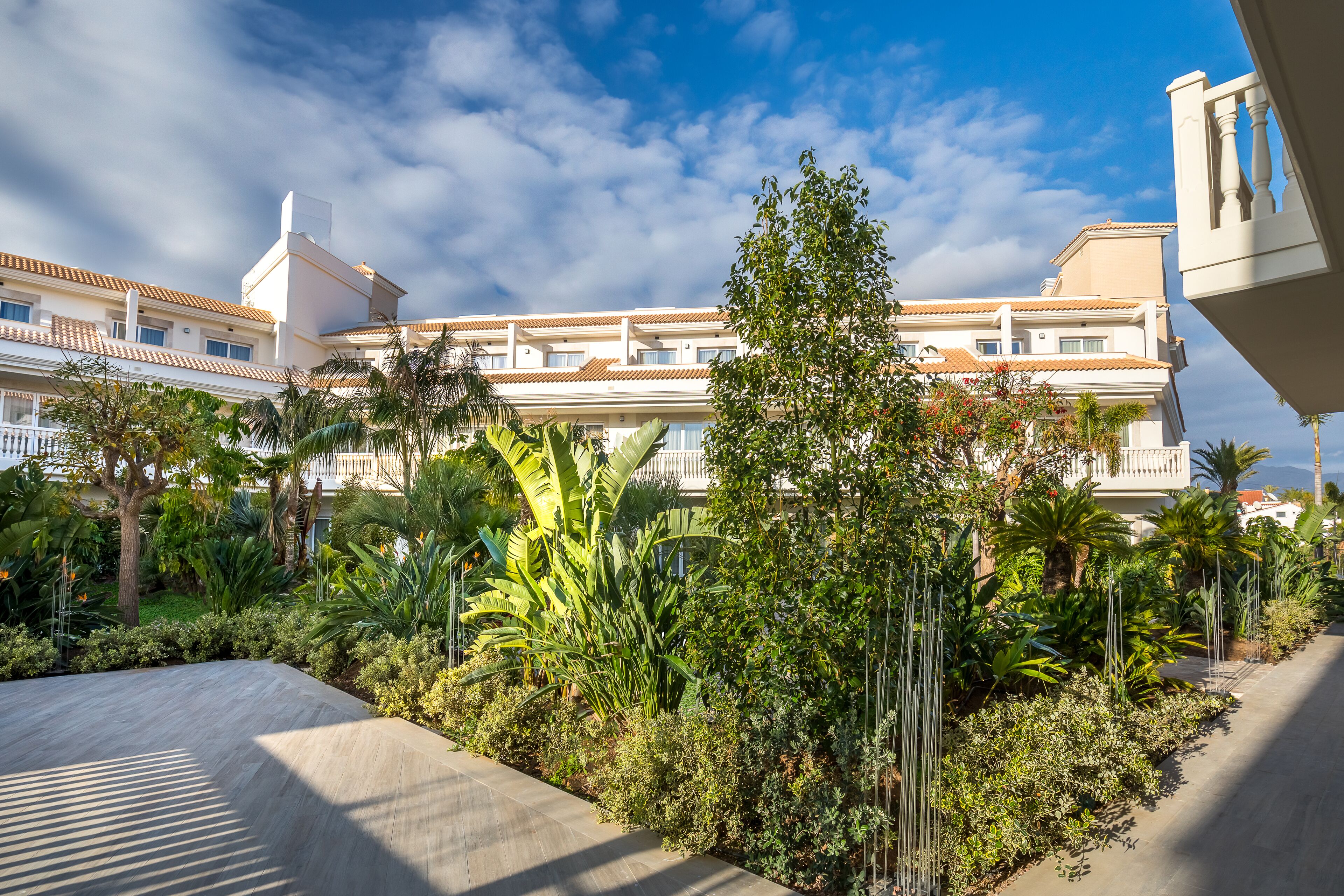 A hotel courtyard with a paved walkway, lush greenery, and a building with multiple balconies and a tiled roof.