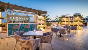 An outdoor dining area with tables and chairs, a bar with a sign reading "BHEAVEN," and a backdrop of plants and a mountain view. - Barcelo La Nucía Hills (La Nucia)