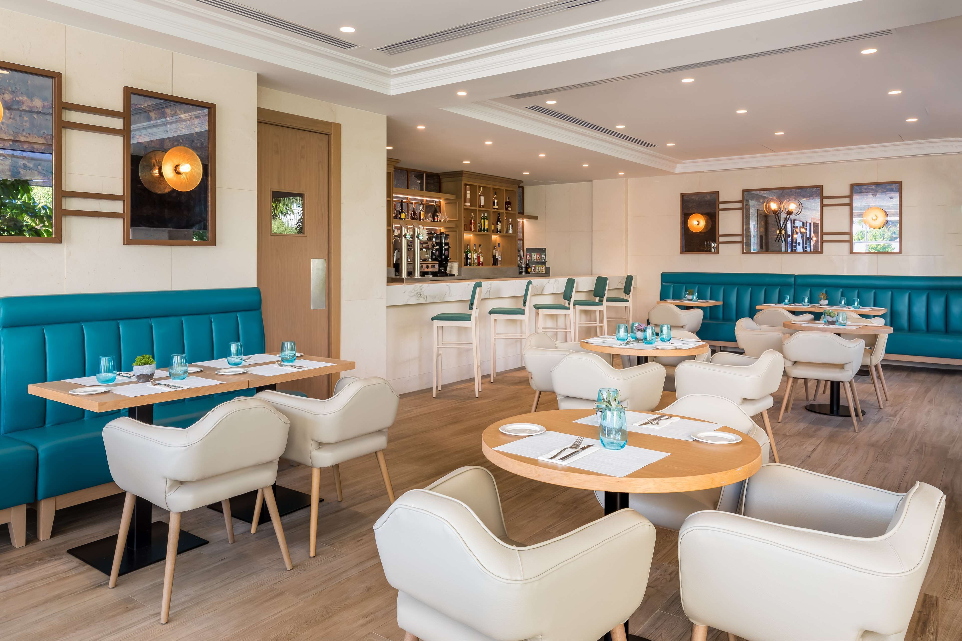 Un restaurant moderne avec des banquettes turquoise, des chaises blanches et des tables rondes. Il y a un coin bar avec des étagères remplies de bouteilles et de verres.