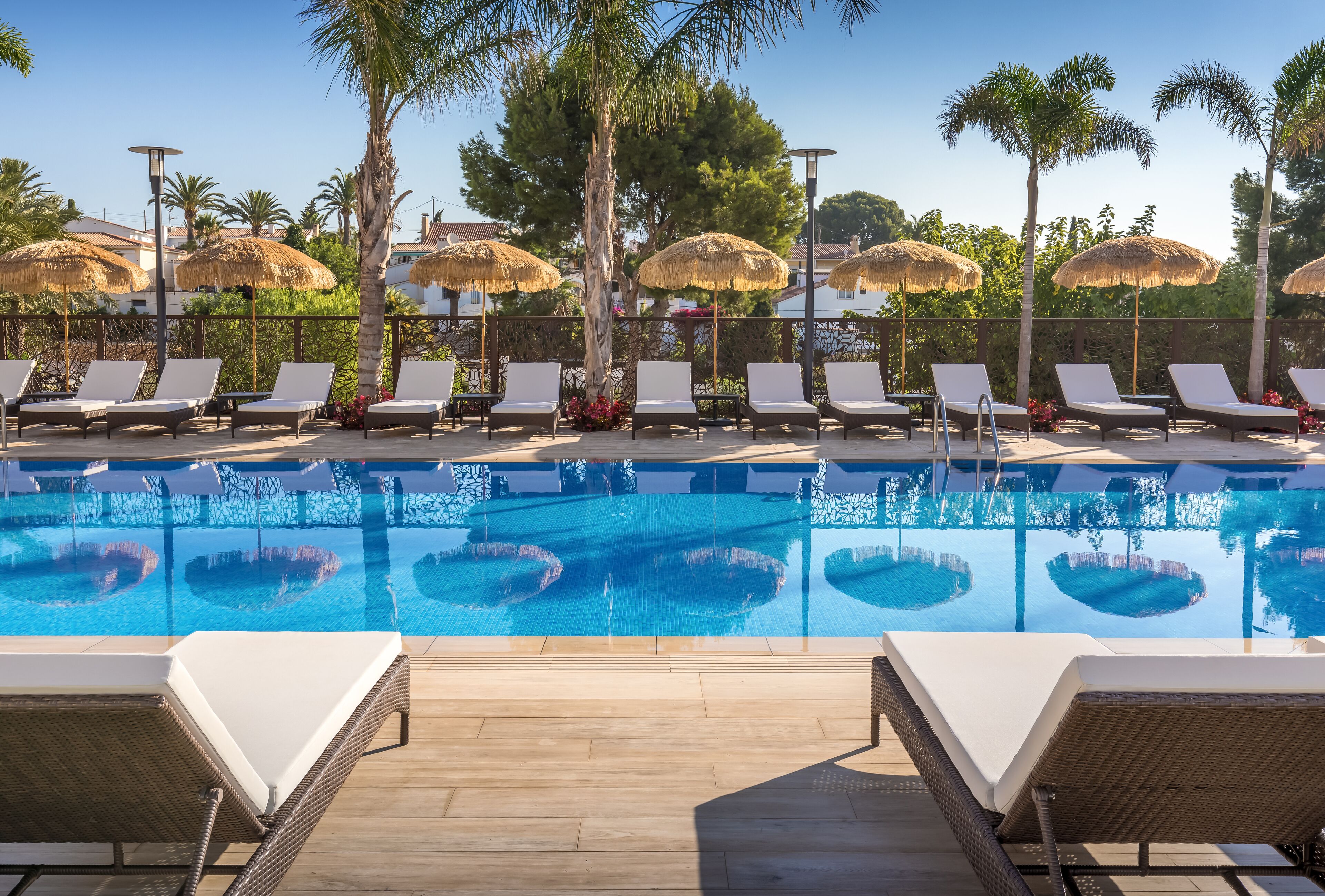 Un área junto a la piscina con sillones de descanso y sombrillas de techo de paja, rodeada de palmeras y una piscina azul cristalina.