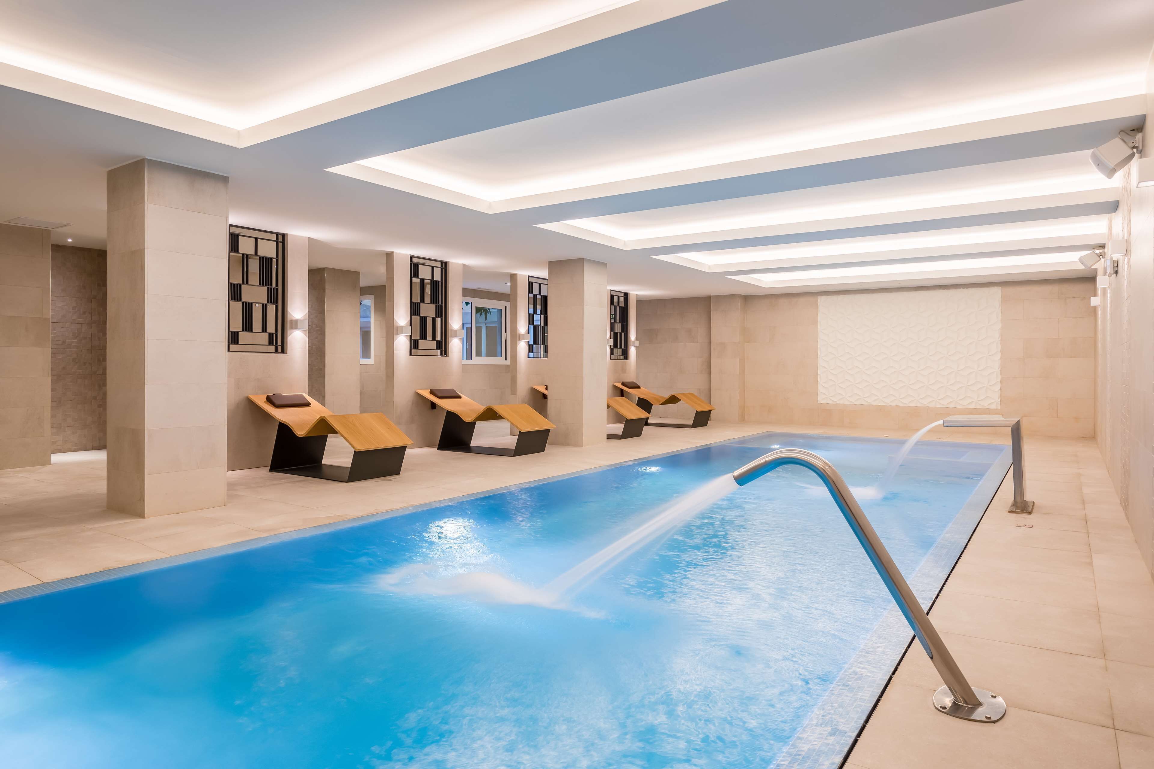 Un espace piscine intérieur moderne, doté d’une longue piscine, de chaises longues et d’un plafond avec des luminaires encastrés.