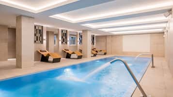 Un espace piscine intérieur moderne, doté d’une longue piscine, de chaises longues et d’un plafond avec des luminaires encastrés.