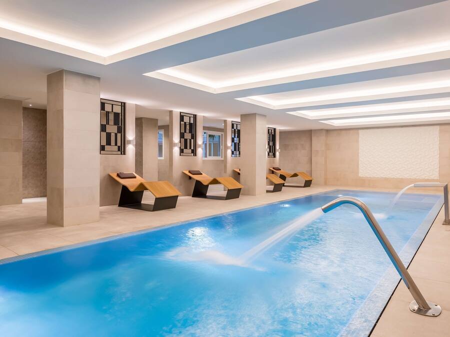 Un espace piscine intérieur moderne, doté d’une longue piscine, de chaises longues et d’un plafond avec des luminaires encastrés.