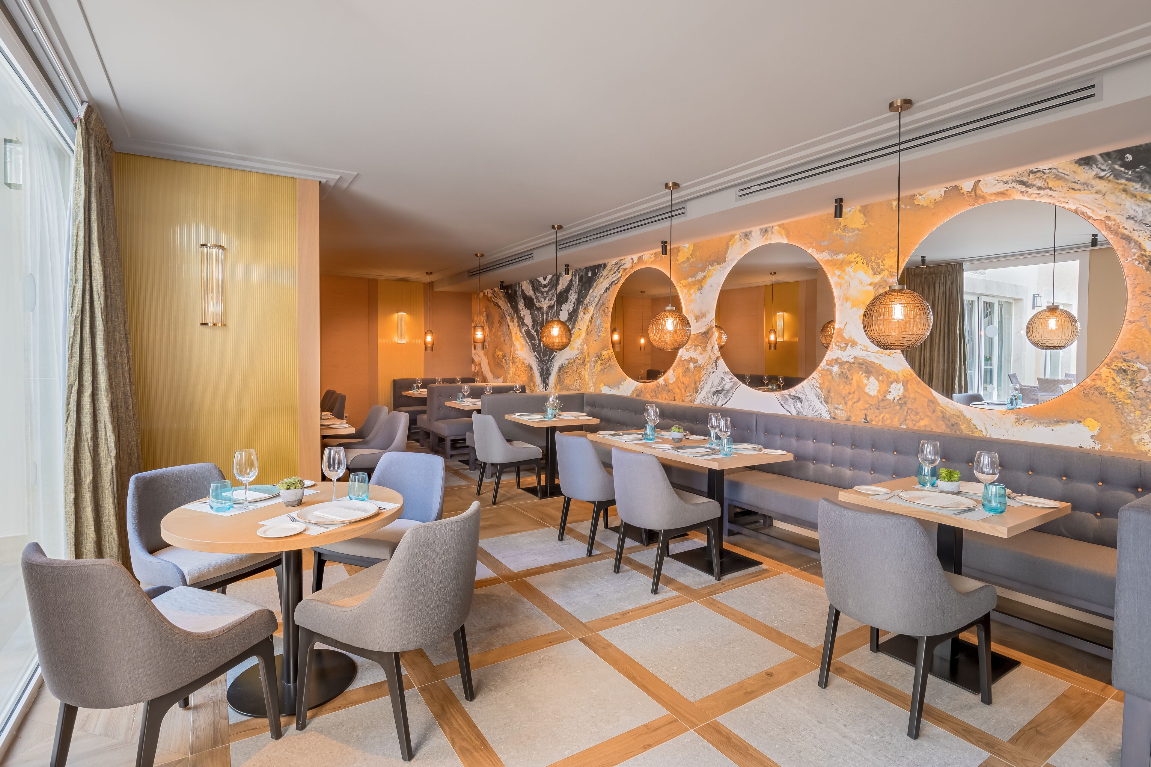 Un restaurante moderno con mesas redondas, sillas grises y una pared de mármol con espejos circulares.