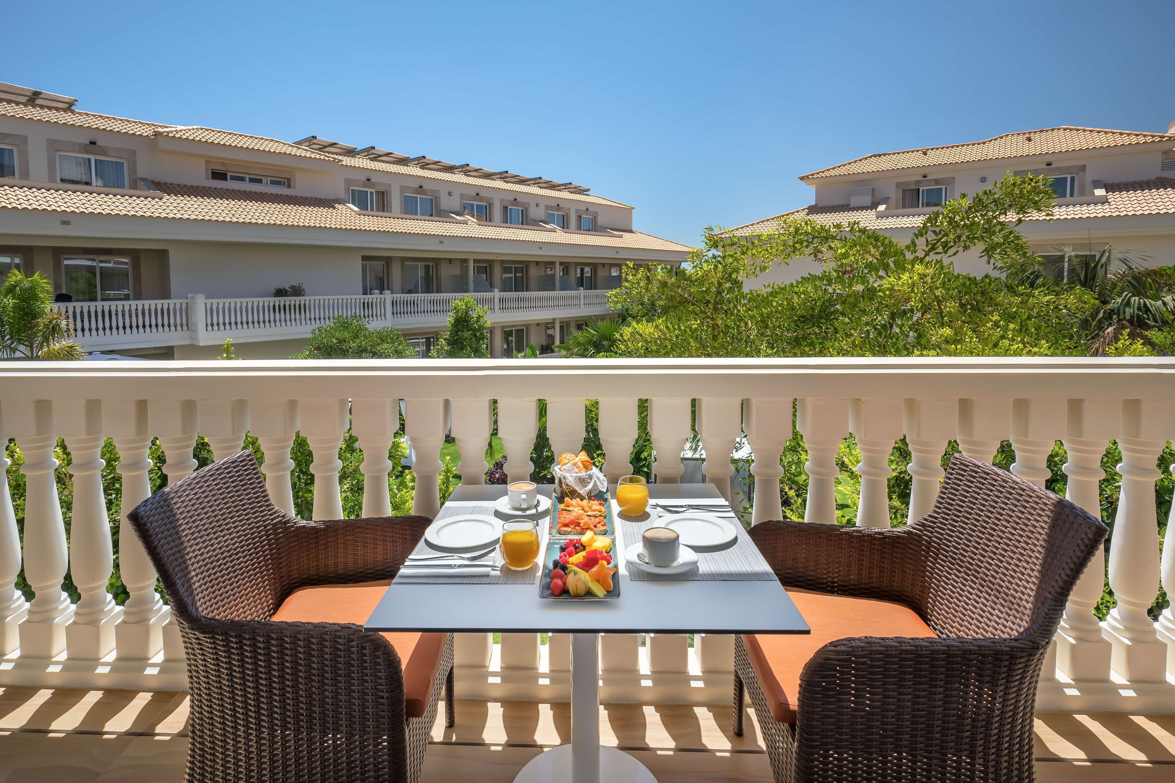A dining table set with breakfast items on a balcony with white balustrades and a view of hotel buildings.